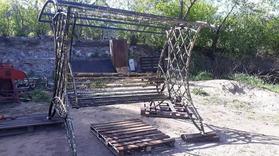 Metalowa huśtawka ogrodowa z ażurowym oparciem i zadaszeniem, umieszczona na paletach w ogrodzie, widoczna w dziennym świetle, z elementami dekoracyjnymi.