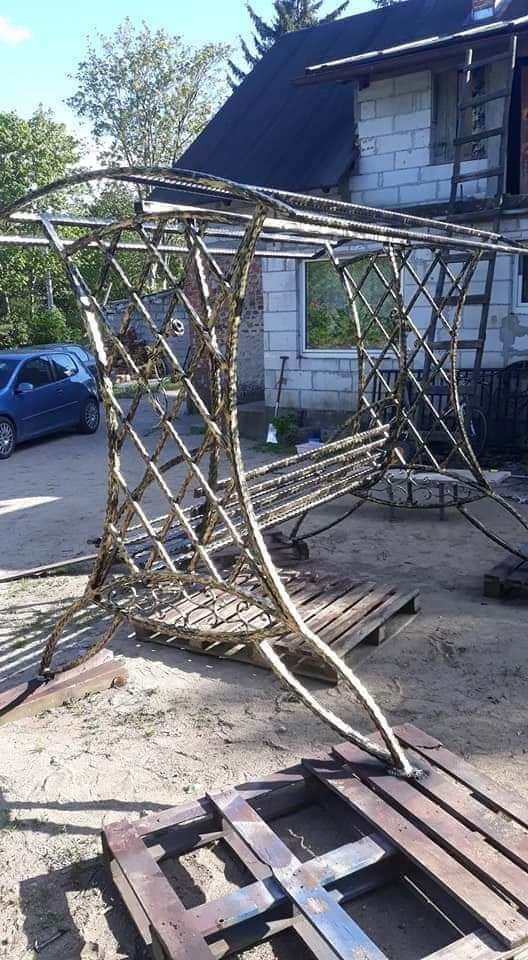 Metalowa konstrukcja w stylu retro, prawdopodobnie altana lub element dekoracyjny, w trakcie budowy na placu budowy, oparta na drewnianych paletach, z widocznym spawem i siatkowym wzorem.