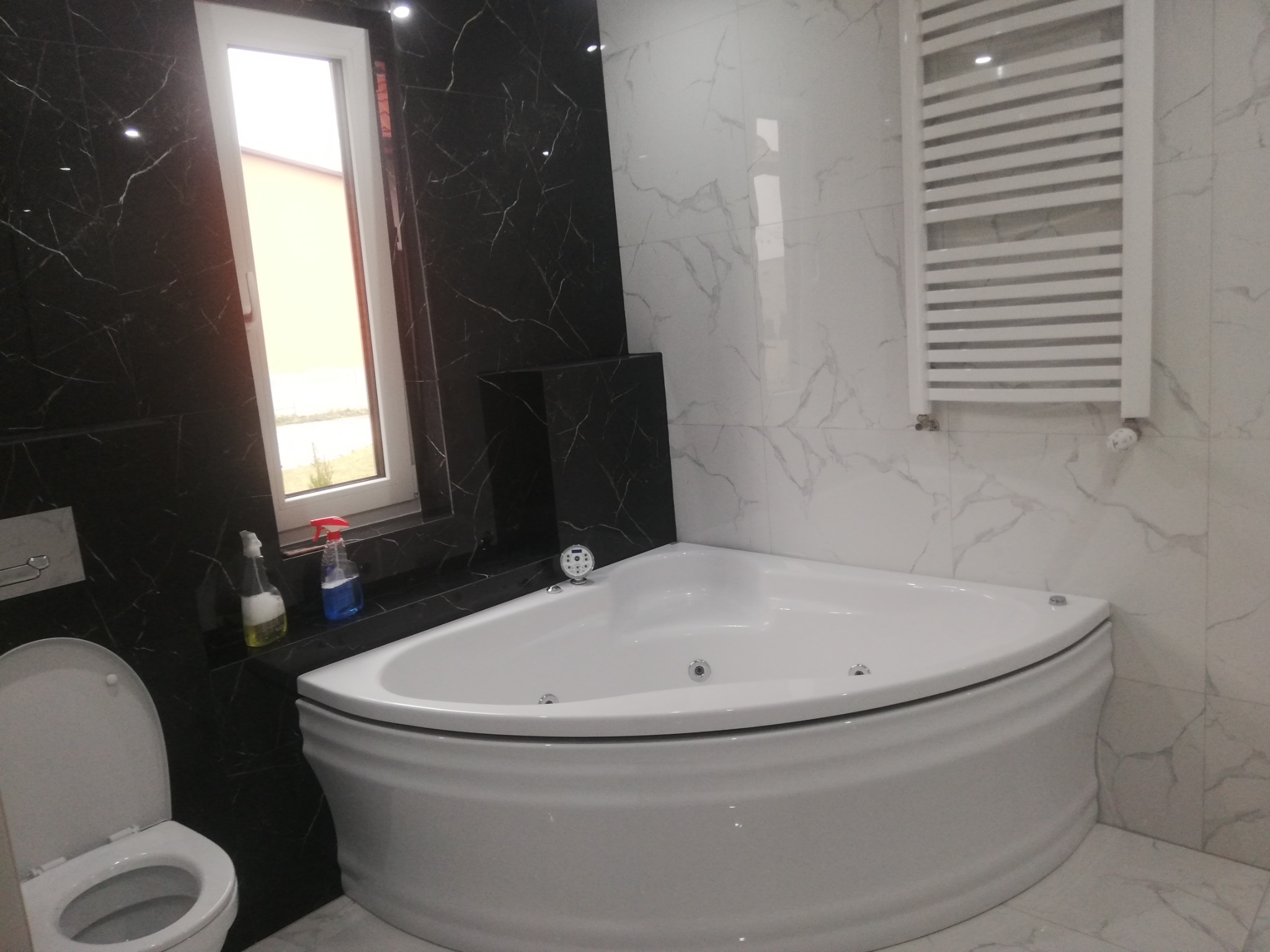 Narożna wanna z hydromasażem w nowoczesnej łazience z czarno-białymi marmurkowymi płytkami, widoczny grzejnik drabinkowy i toaleta.