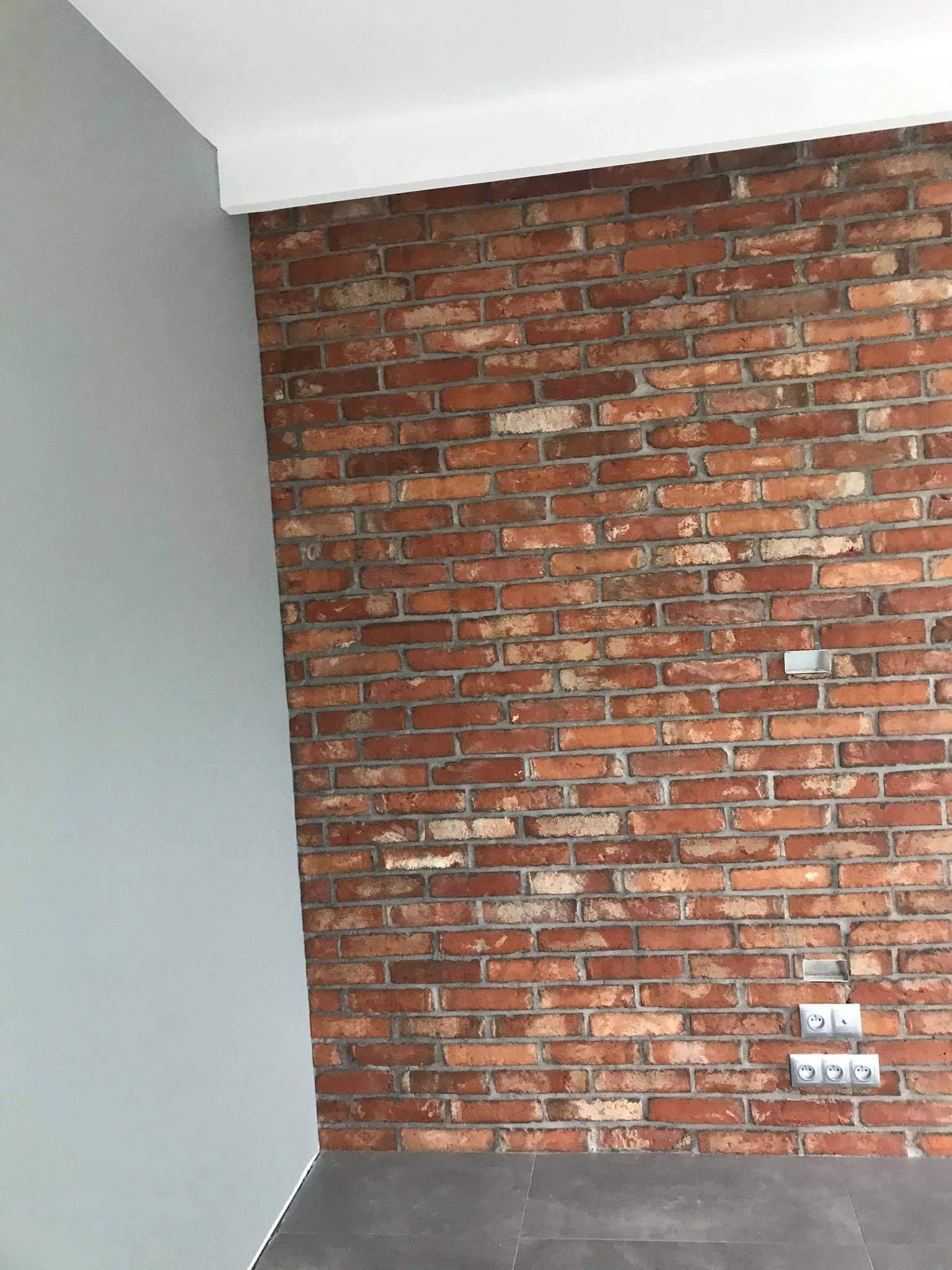 Ściana z czerwonej cegły z widocznymi gniazdkami elektrycznymi, obok szara ściana i biały sufit, szara podłoga.