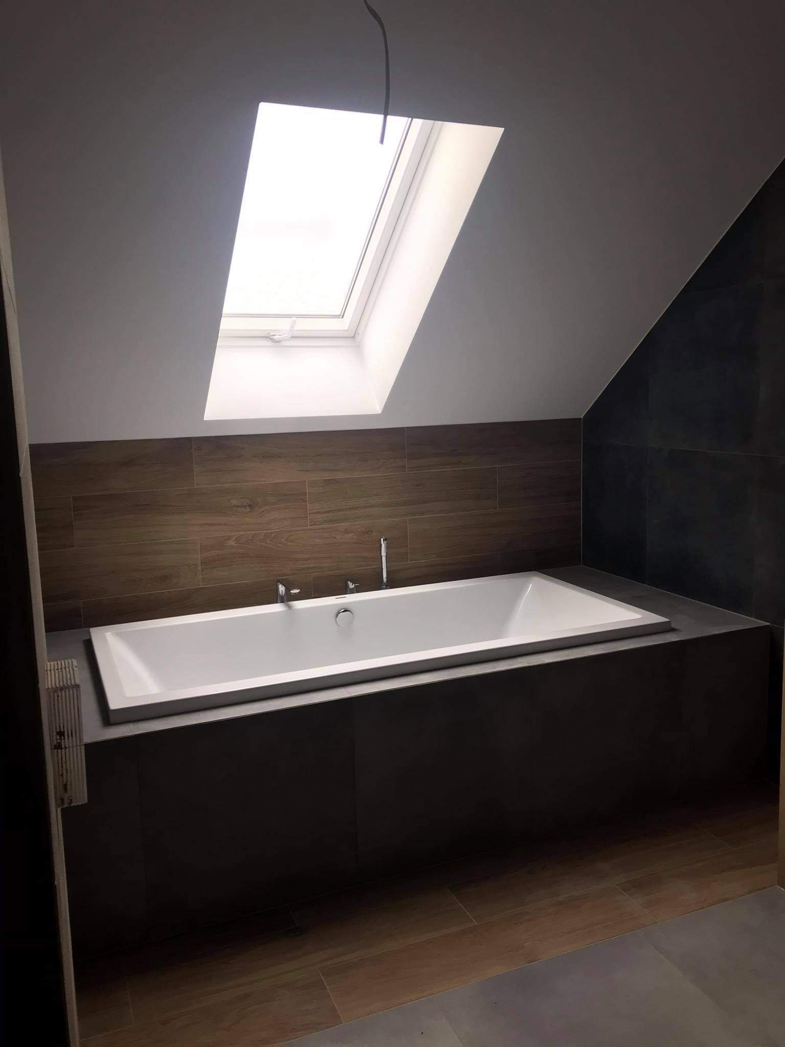 Nowoczesna wanna w zabudowie, obłożona ciemnymi płytkami, z armaturą i oknem dachowym w łazience na poddaszu.