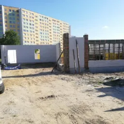 Wznoszenie ścian parterowego domu z bloczków w Szczercowskiej Wsi, widoczne szalunki okienne i zbrojenia, na pierwszym planie piasek i zaparkowany samochód dostawczy.