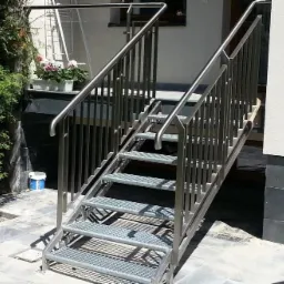 Zewnętrzne schody z metalową kratownicą i stalową balustradą, prowadzące do budynku z oknem i kwiatami w doniczkach. Na pierwszym planie widoczny fragment białego krzesła.