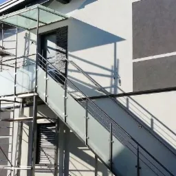 Zewnętrzne schody z balustradami ze szkła matowego i metalu prowadzące do wejścia na piętro budynku w Sieprawiu, widoczne rusztowanie po lewej stronie.