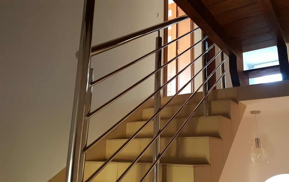 Schody wykończone beżowymi płytkami z nowoczesną balustradą ze stali nierdzewnej z poziomymi prętami, widok od dołu, w tle drewniane elementy konstrukcyjne i drzwi.