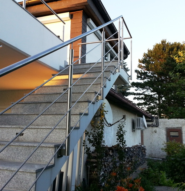 Zewnętrzne schody z szarego kamienia z prostą, metalową balustradą z rurek, prowadzące do wejścia na piętro domu.
