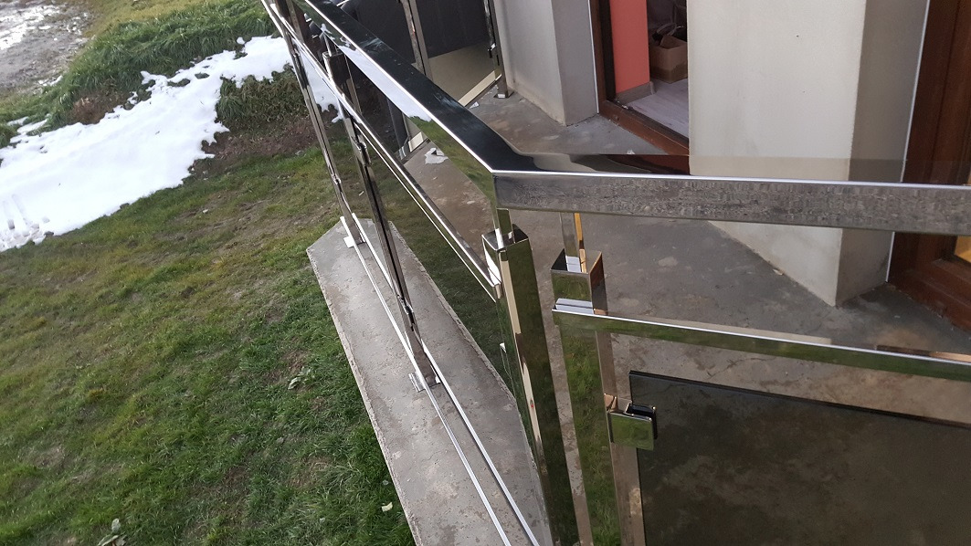 Szklana balustrada balkonowa z elementami ze stali nierdzewnej, widok z góry na trawnik z resztkami śniegu.