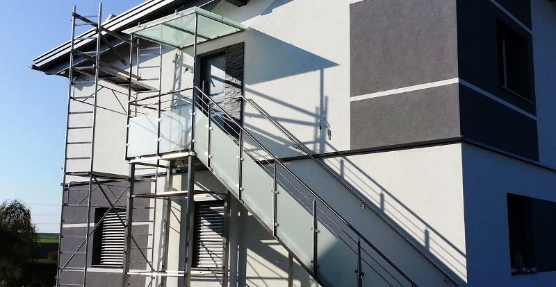 Zewnętrzne schody z balustradami ze szkła matowego i metalu prowadzące do wejścia na piętro budynku w Sieprawiu, widoczne rusztowanie po lewej stronie.