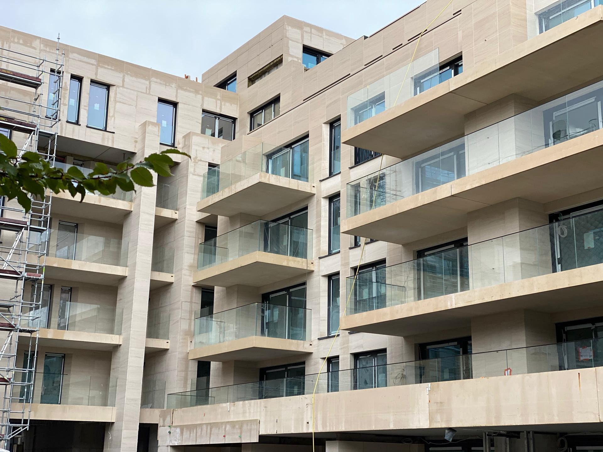 Wielopiętrowy budynek z balkonami zabezpieczonymi szkłem, w trakcie budowy, widoczne rusztowania po lewej stronie.