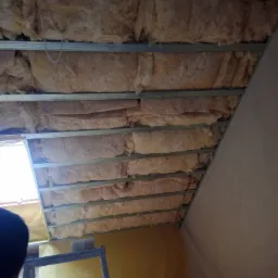 Skośny sufit poddasza z izolacją z wełny mineralnej, widoczny metalowy stelaż i fragment drabiny malarskiej.