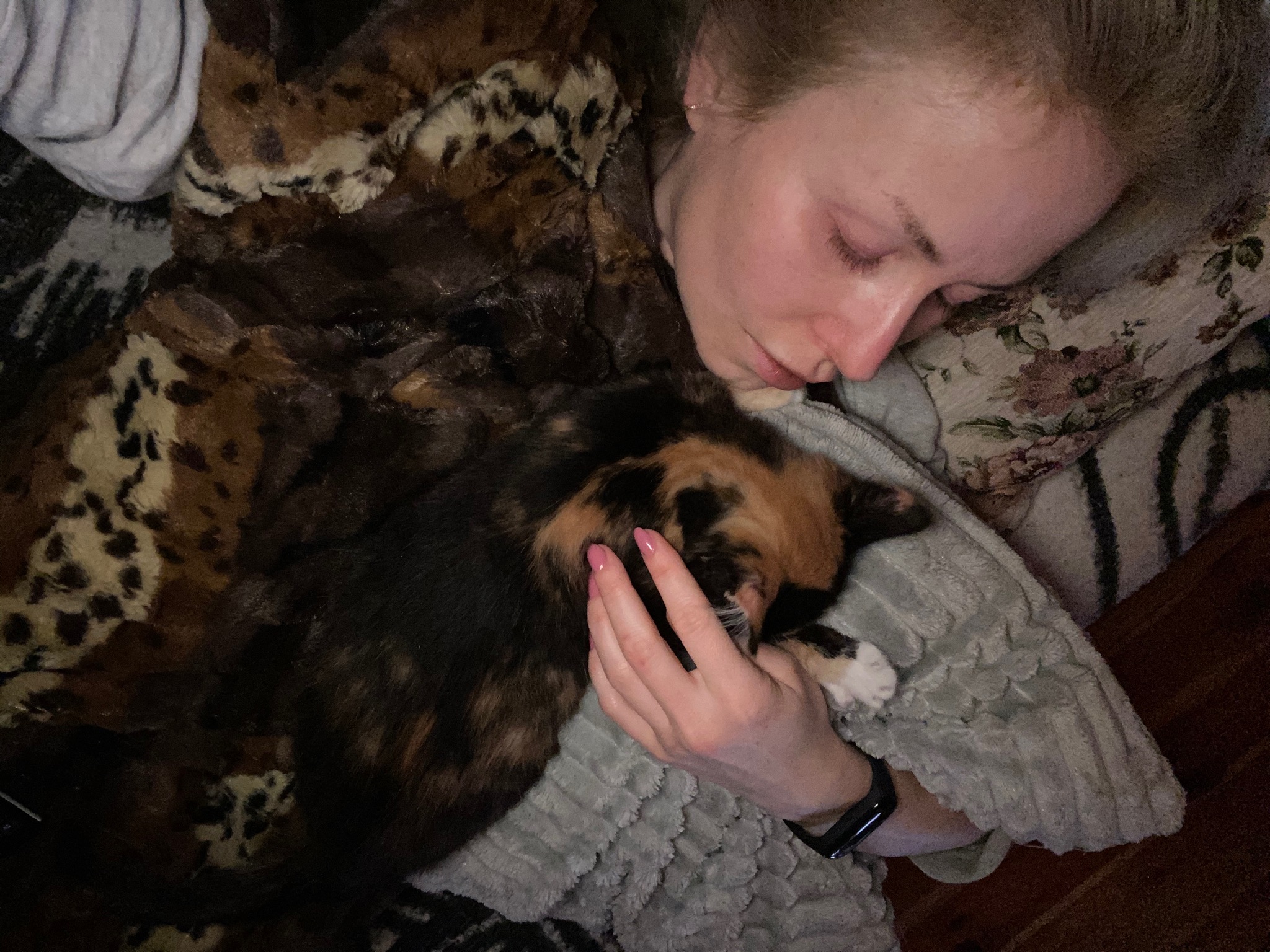 Śpiąca kobieta z trójkolorowym kotem, wtulona w futrzaną narzutę w panterkę, na poduszce w kwiaty.