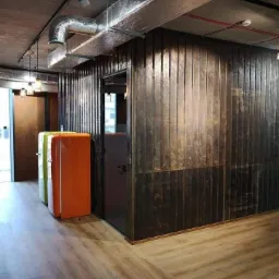 Biuro z drewnianą ścianą, szklanymi drzwiami, widocznymi instalacjami wentylacyjnymi pod sufitem oraz kolorowymi lodówkami w stylu retro.