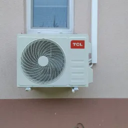 Zewnętrzna jednostka klimatyzacji TCL zamontowana na dwukolorowej, otynkowanej ścianie budynku, widoczny fragment okna i rury odprowadzającej skropliny.