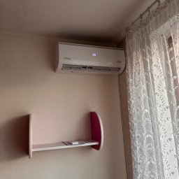 INSTAL4U - Biały klimatyzator na ścianie w pokoju, obok półka i firanka. Widoczny wyświetlacz z temperaturą 26 stopni. Ujęcie z bliska, wnętrze mieszkania.