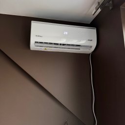 INSTAL4U - Biały klimatyzator Aura Line z wyświetlaczem 28 stopni Celsjusza, zamontowany na brązowej ścianie z widocznym kablem zasilającym i fragmentem szyny sufitowej.
