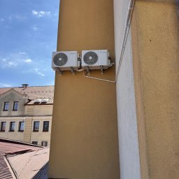 INSTAL4U - Dwa białe klimatyzatory zamontowane na żółtej ścianie budynku, widoczne rury instalacyjne i fragment dachu sąsiedniego budynku na tle błękitnego nieba.