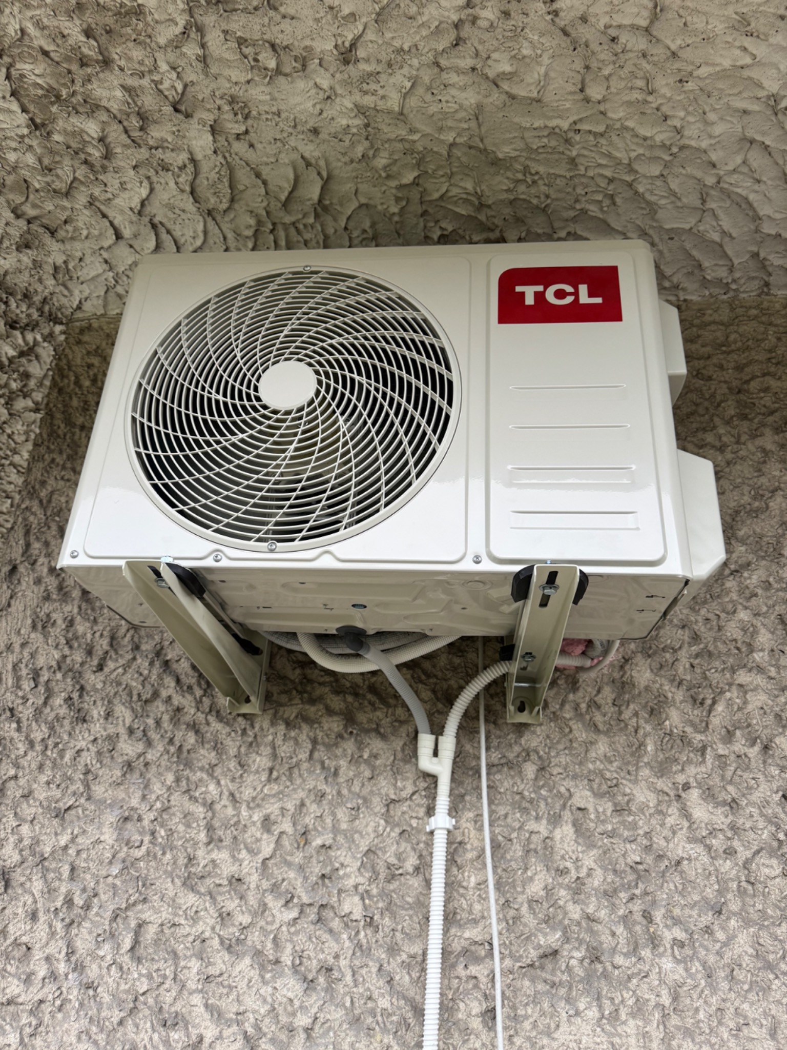 Biała jednostka zewnętrzna klimatyzatora TCL zamontowana na szarej, chropowatej ścianie zewnętrznej budynku. Widoczne białe przewody i metalowe wsporniki montażowe.