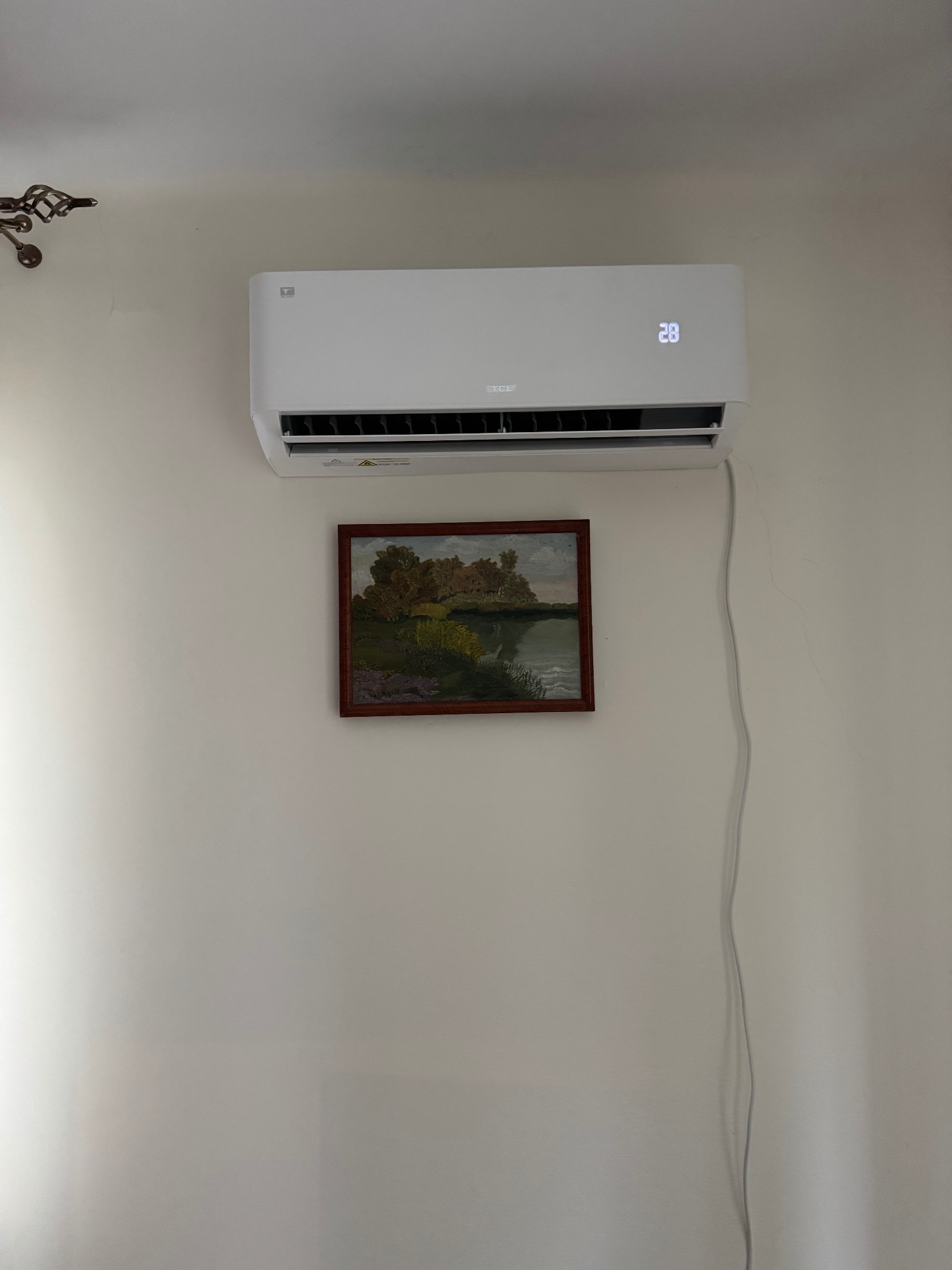 Biały klimatyzator ścienny marki TCL, z wyświetlaczem wskazującym temperaturę 28 stopni Celsjusza, zamontowany nad obrazem z motywem krajobrazu w brązowej ramie, na jasnej ścianie.