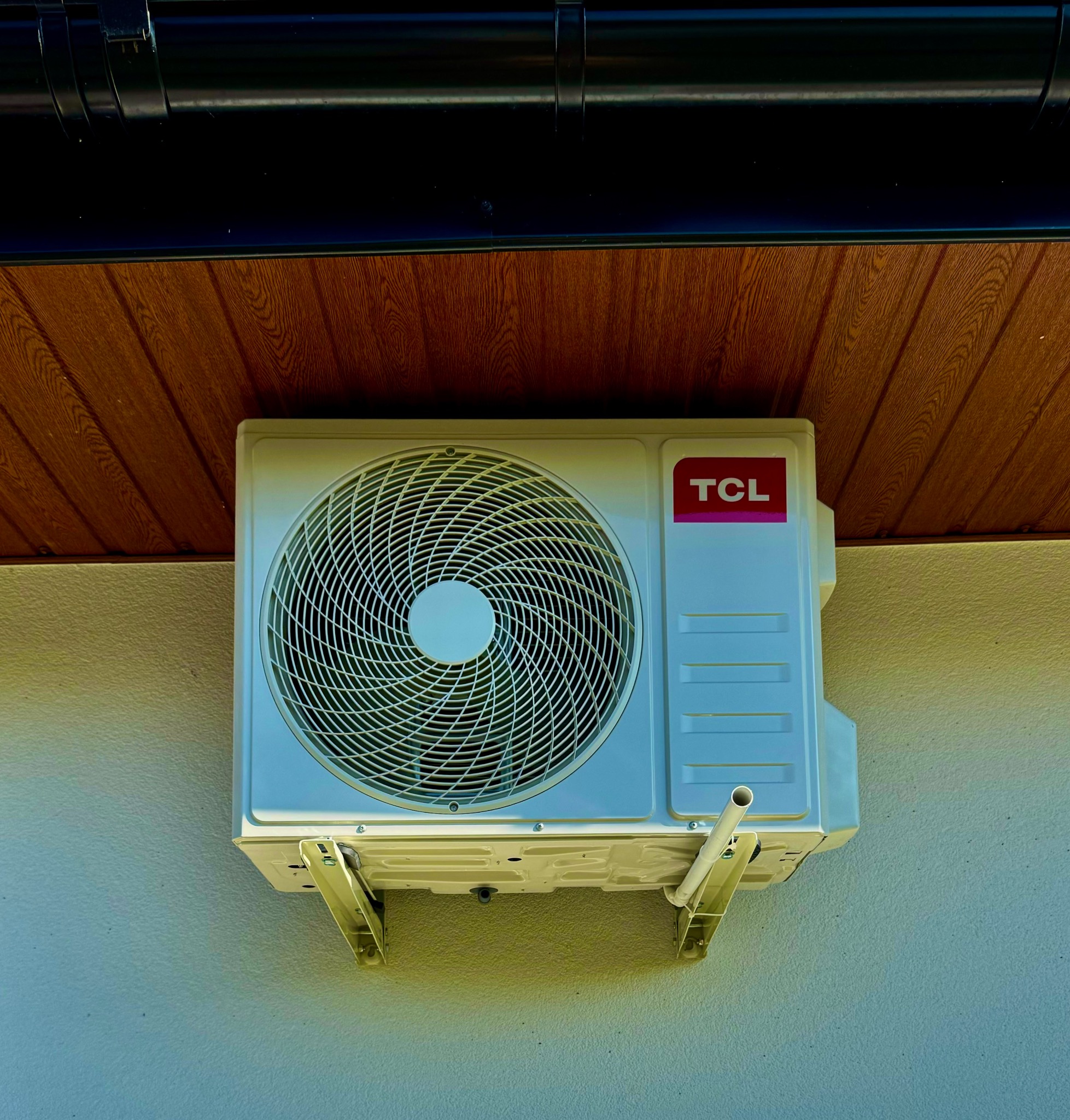 Zewnętrzna jednostka klimatyzacji TCL zamontowana na elewacji budynku, widoczny odpływ skroplin i brązowa podbitka dachowa.