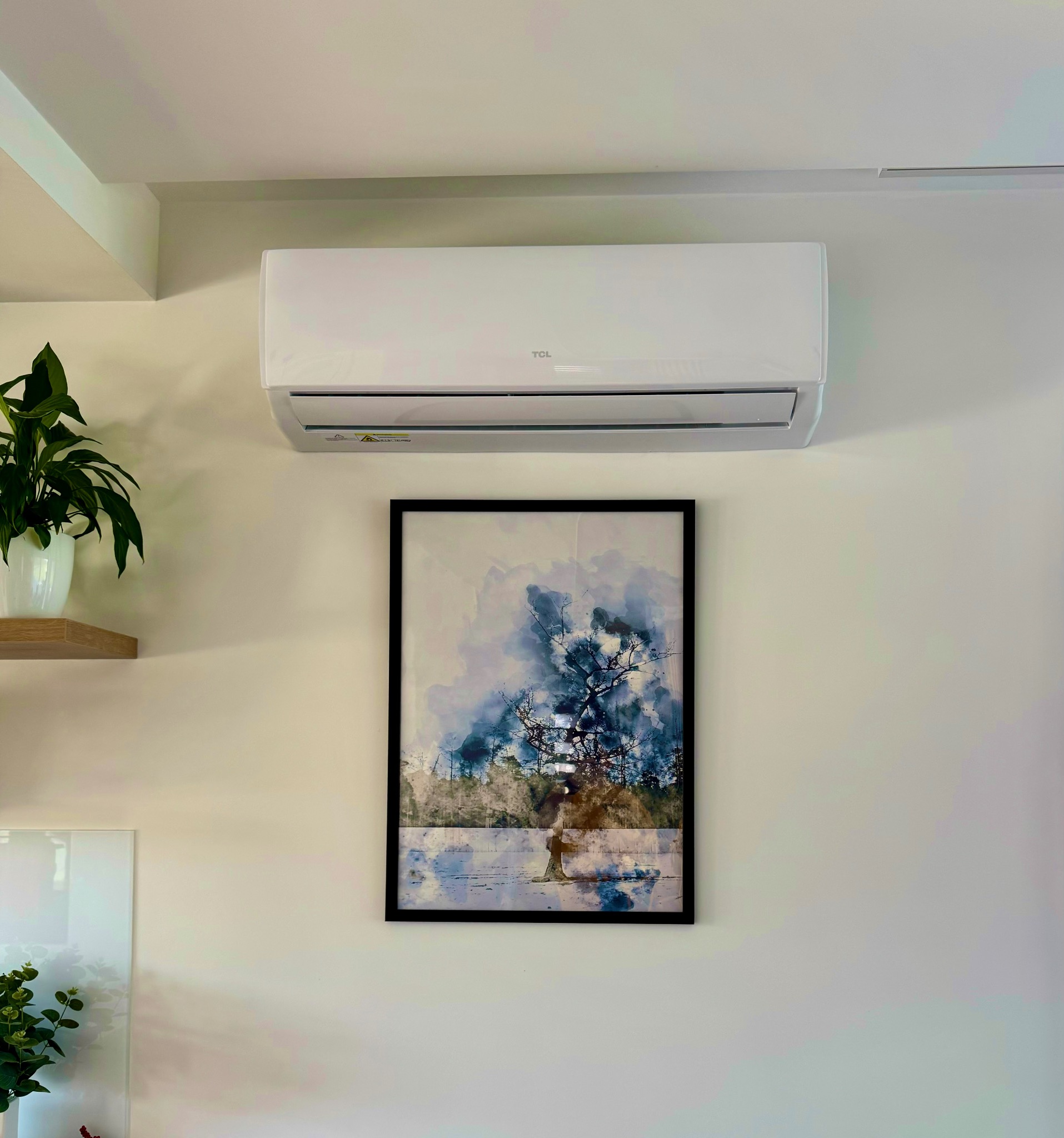 Biały klimatyzator TCL zamontowany na ścianie nad obrazem w czarnej ramie, obok półki z rośliną doniczkową.