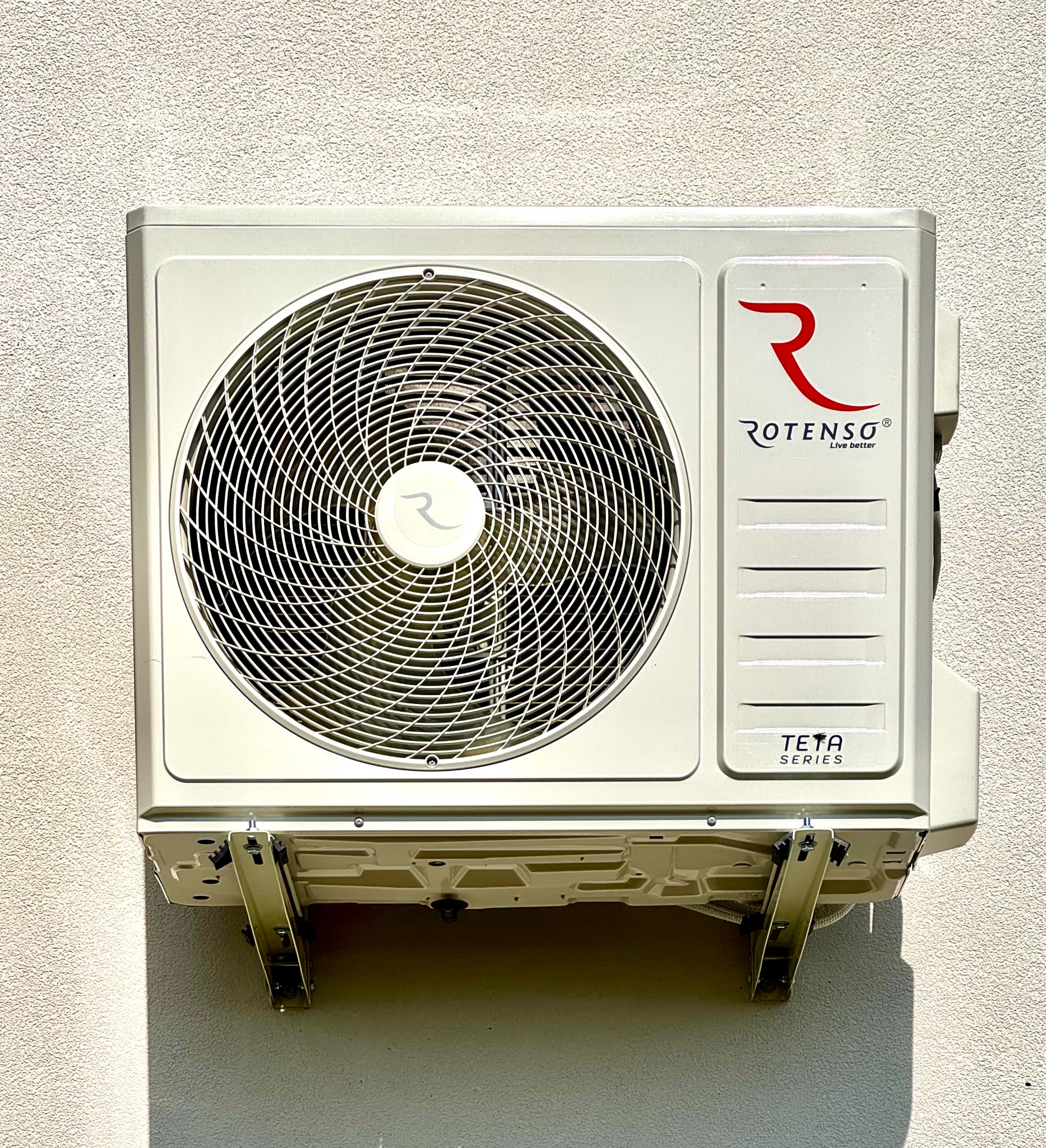 Zewnętrzna jednostka klimatyzacji Rotenso Teta Series zamontowana na ścianie o jasnej, chropowatej fakturze, widoczne wsporniki montażowe i cień rzucany przez urządzenie.