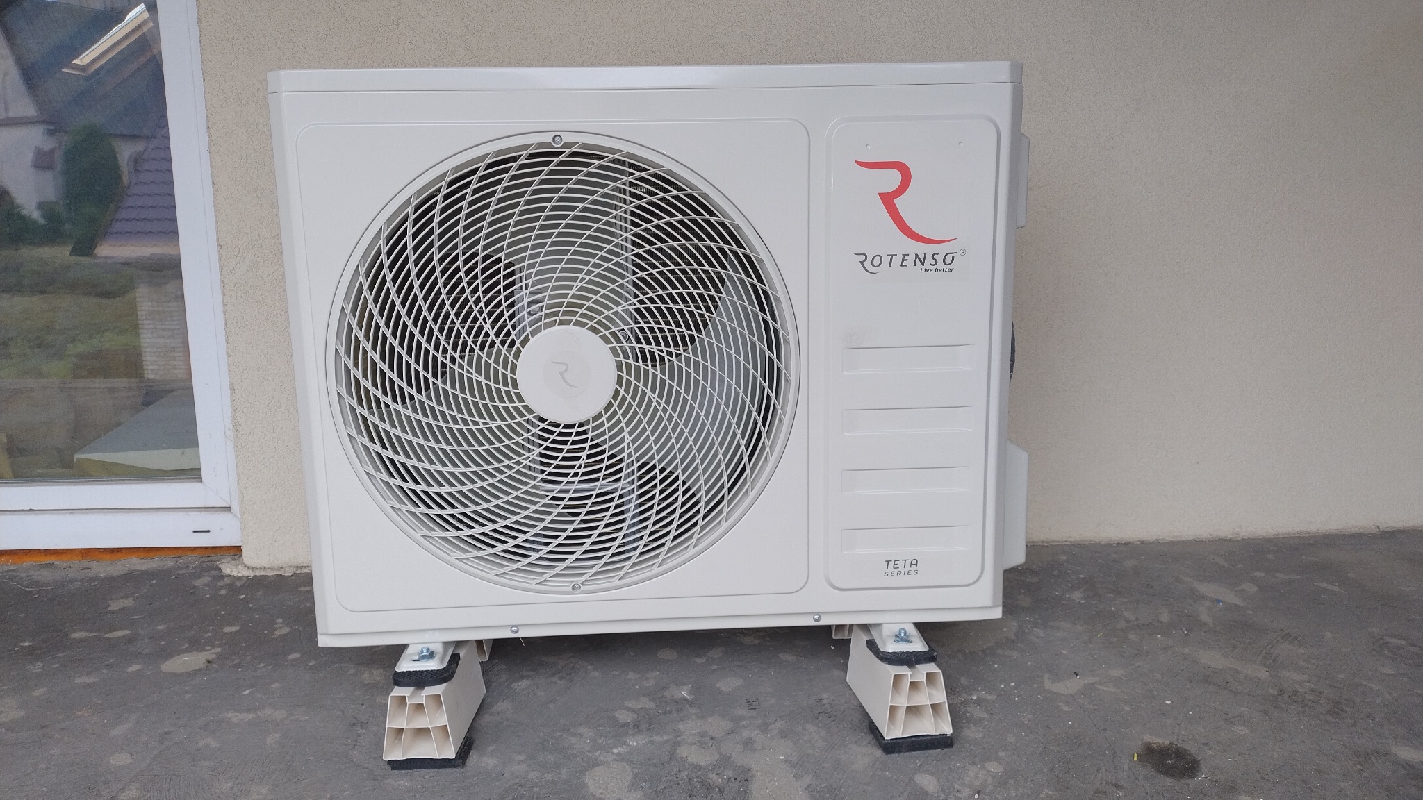 Zewnętrzna jednostka klimatyzacji Rotenso TETA Series, zamontowana na specjalnych wspornikach z tworzywa sztucznego przy ścianie budynku z widocznym oknem.