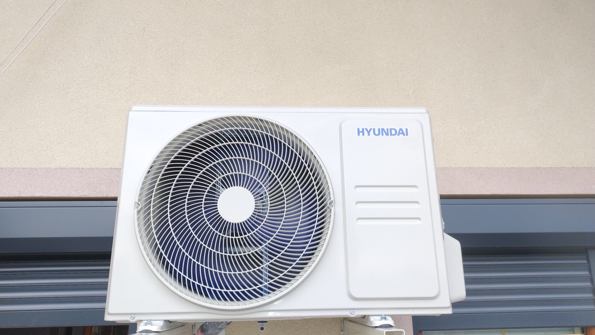 Zewnętrzna jednostka klimatyzacji Hyundai zamontowana na ścianie budynku z widoczną roletą okienną w tle.