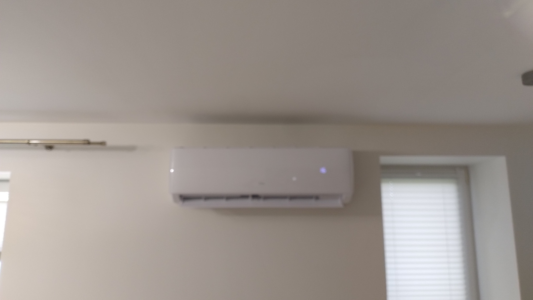 Biały klimatyzator ścienny zamontowany w jasnym pokoju, widoczne żaluzje okienne w tle.