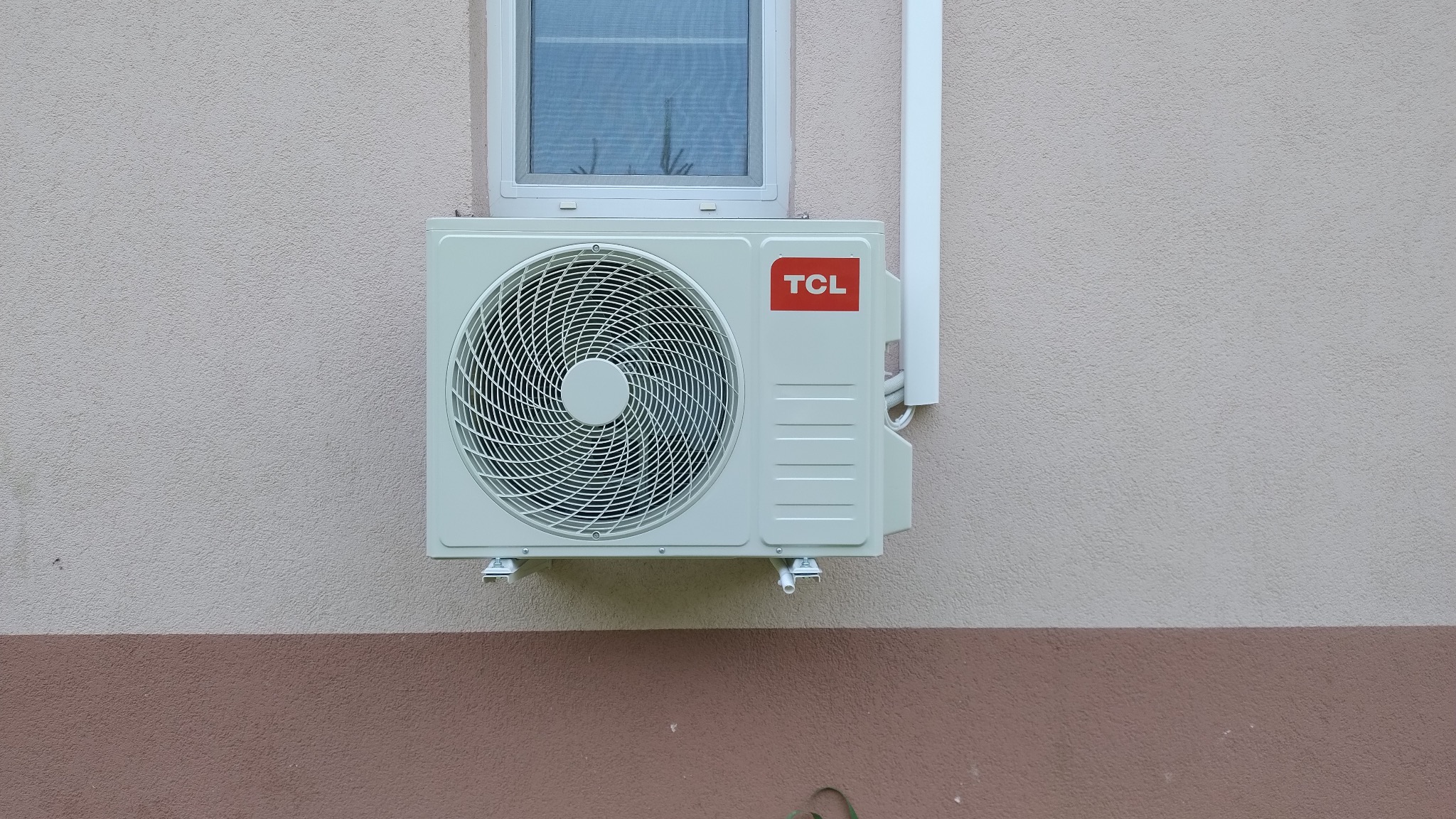 Zewnętrzna jednostka klimatyzacji TCL zamontowana na dwukolorowej, otynkowanej ścianie budynku, widoczny fragment okna i rury odprowadzającej skropliny.