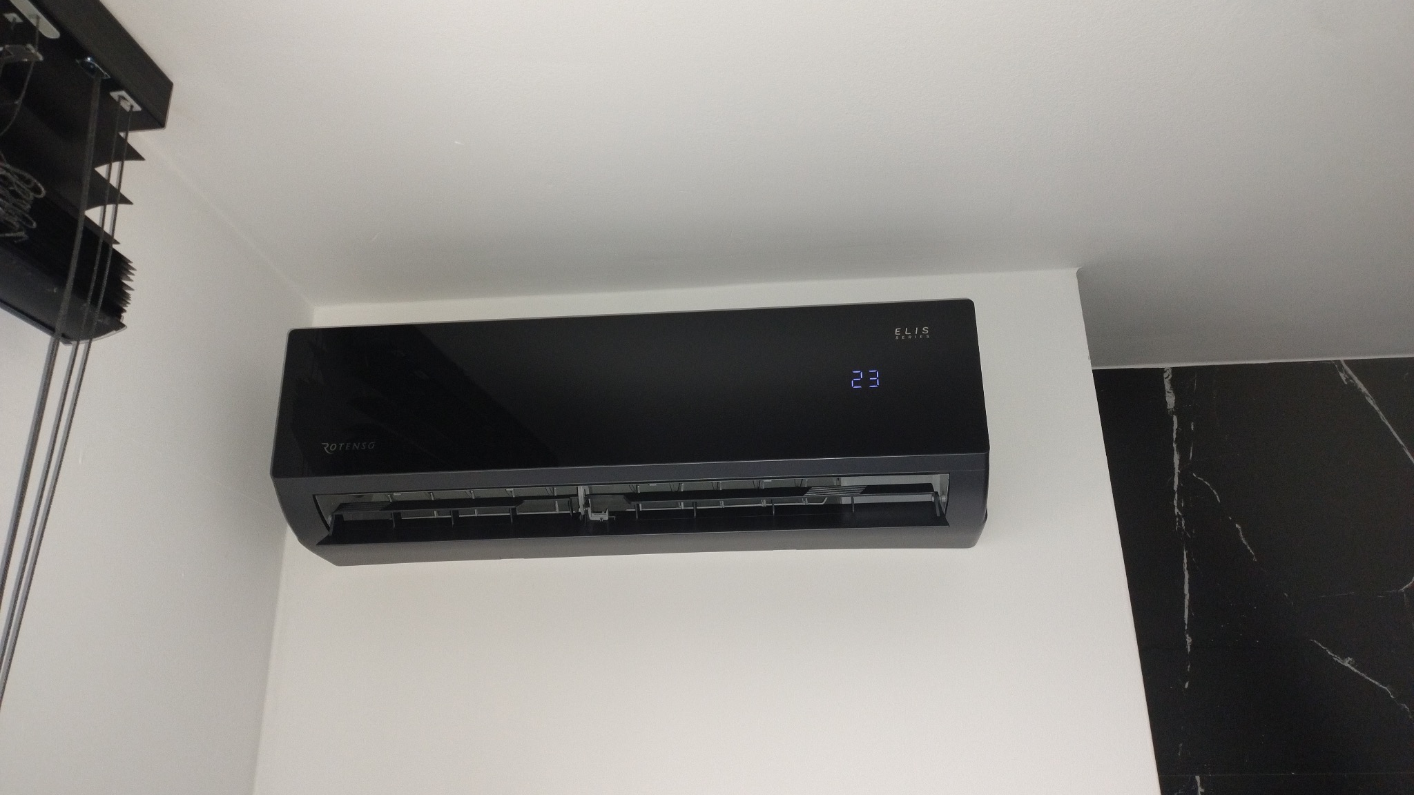 Czarny klimatyzator Rotenso Elis Series z wyświetlaną temperaturą 23 stopnie, zamontowany na białej ścianie, z fragmentem czarnych marmurowych płytek w tle.
