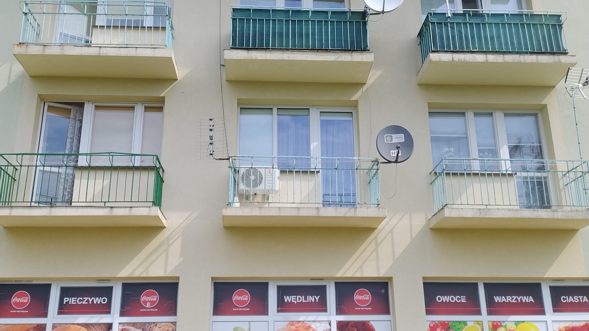 Elewacja budynku z balkonami, na jednym z balkonów zamontowana jednostka zewnętrzna klimatyzacji typu split, obok antena satelitarna, na dole okna sklepowe z reklamami Coca-Coli.