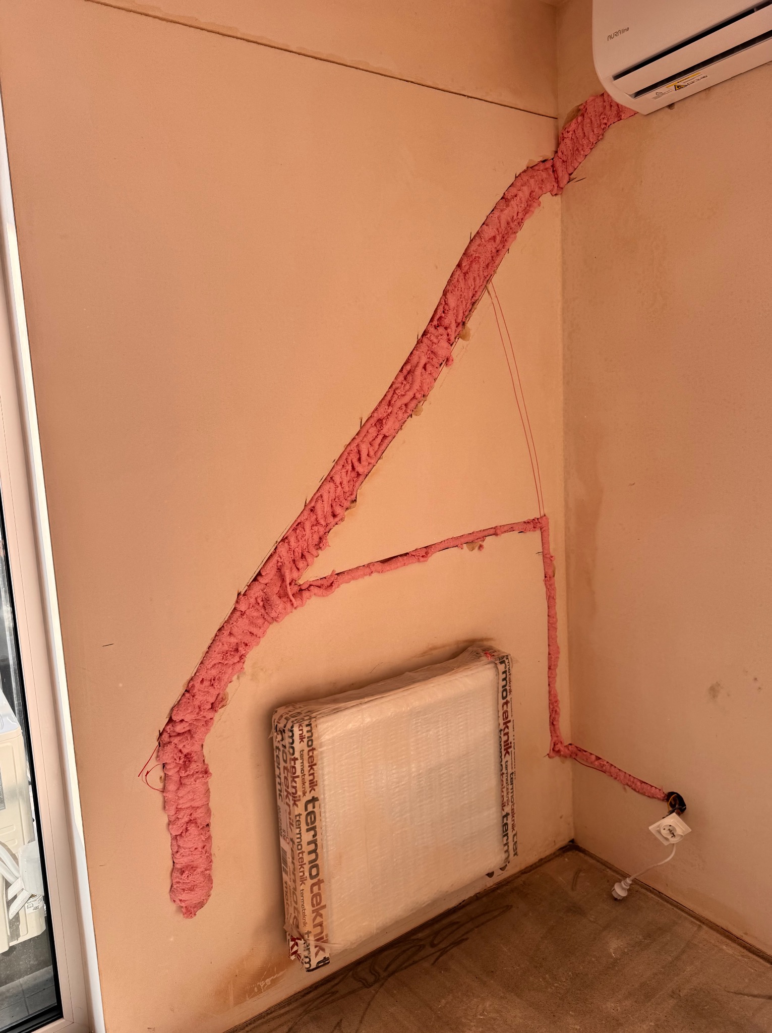 Montaż klimatyzacji: różowa pianka uszczelniająca przewody w ścianie, widoczny klimatyzator Aura Line i grzejnik Termoteknik. Instalacja w trakcie prac.