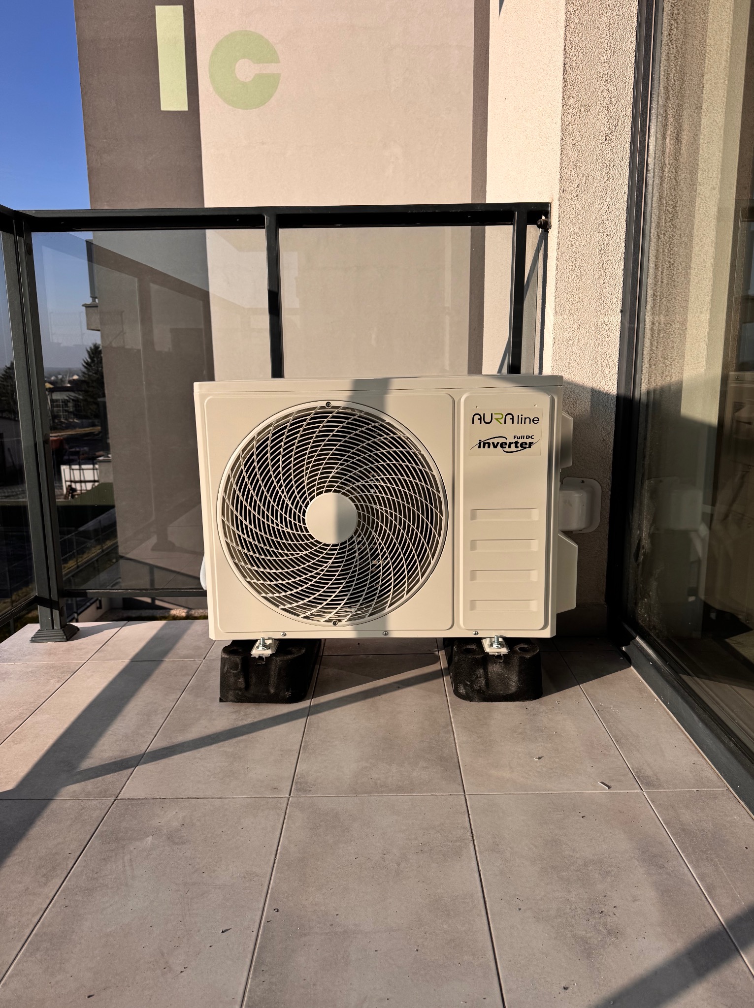 Biały klimatyzator Aura Line Inverter na balkonie, zamontowany na czarnych podkładkach antywibracyjnych. Widok z bliska, zarys miasta w tle.