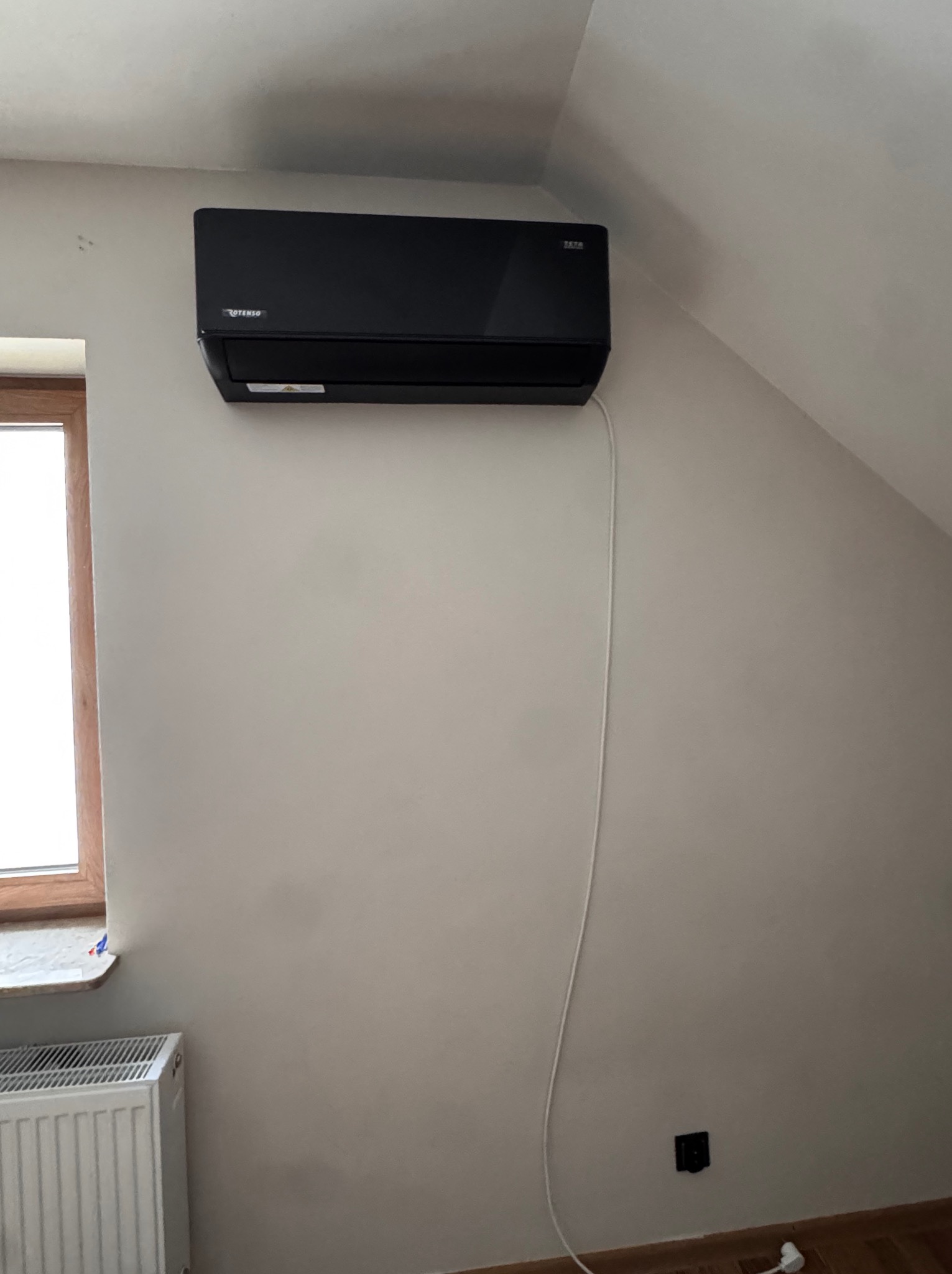 Czarny klimatyzator Rotenso zamontowany na ścianie w pokoju na poddaszu, widoczny kabel zasilający i grzejnik pod oknem. Ściana w kolorze złamanej bieli.