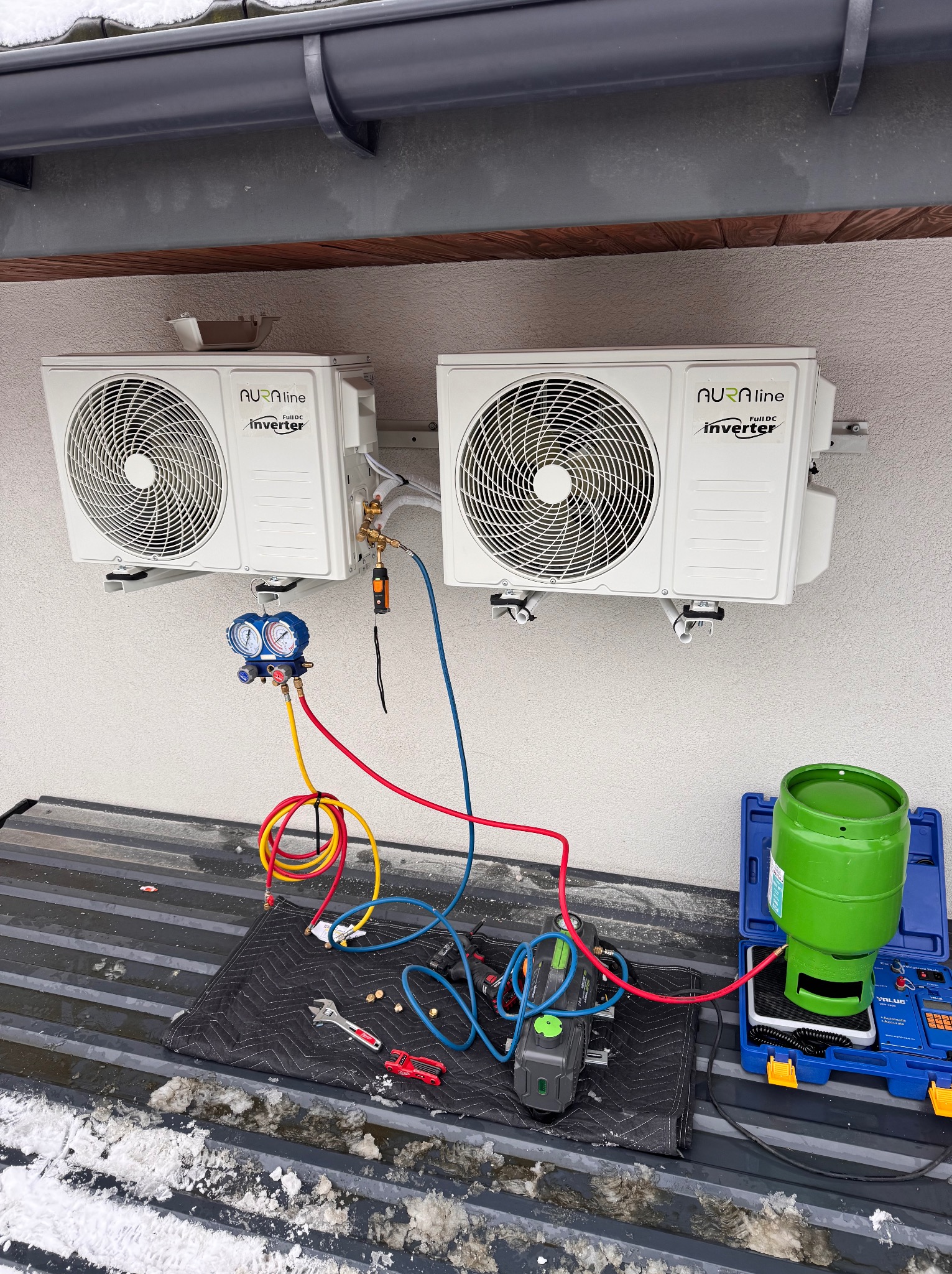 Dwa białe klimatyzatory Aura Line zamontowane na ścianie budynku, podłączone do narzędzi serwisowych, butla z czynnikiem chłodniczym obok. Prace serwisowe w toku.