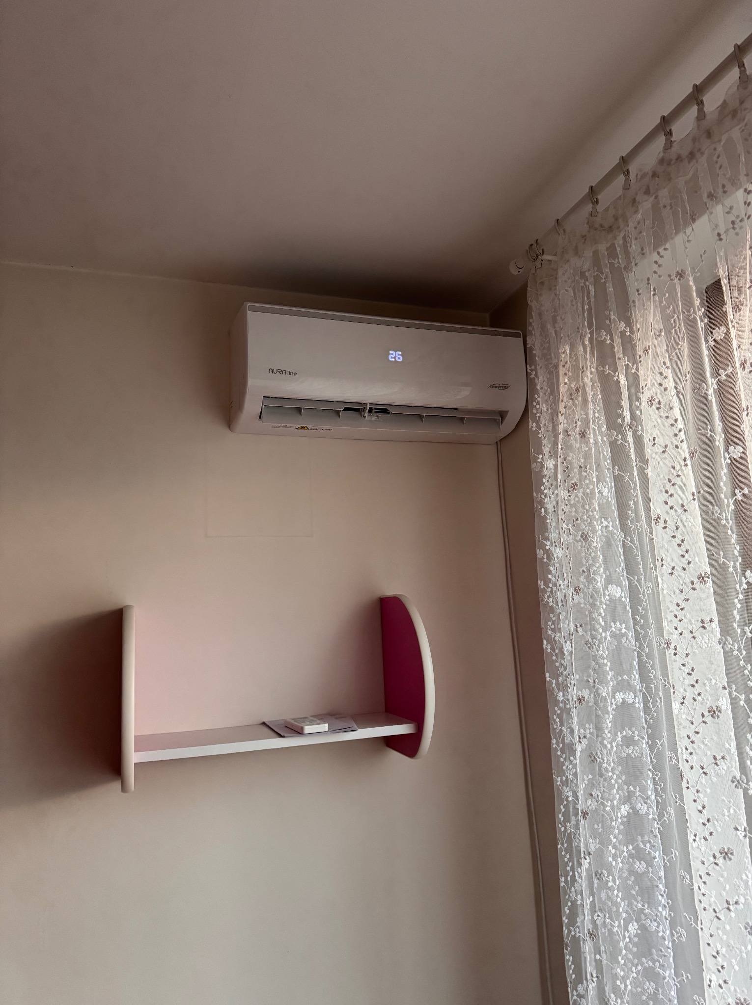 Biały klimatyzator na ścianie w pokoju, obok półka i firanka. Widoczny wyświetlacz z temperaturą 26 stopni. Ujęcie z bliska, wnętrze mieszkania.