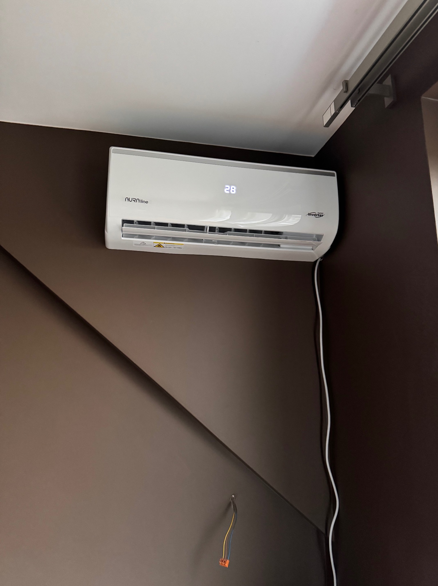 Biały klimatyzator Aura Line z wyświetlaczem 28 stopni Celsjusza, zamontowany na brązowej ścianie z widocznym kablem zasilającym i fragmentem szyny sufitowej.