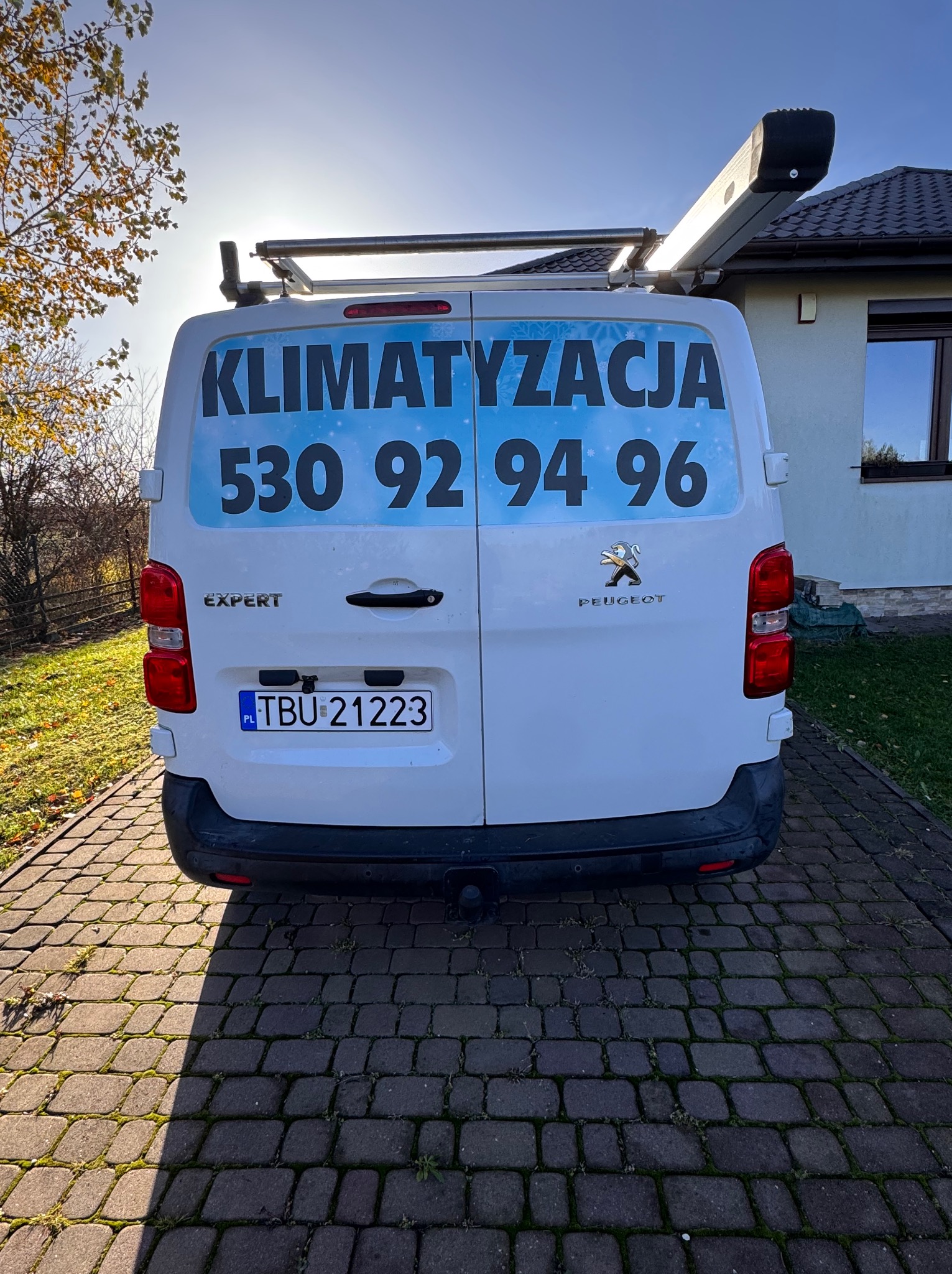 Tył białego samochodu dostawczego Peugeot Expert z reklamą firmy klimatyzacyjnej i numerem telefonu na tle domu i drzew.
