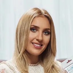 Uśmiechnięta blondynka z niebieskimi oczami, w białej koronkowej bluzce, trzymająca w ręku opakowanie produktu, na rozmytym tle.