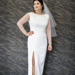 Panna młoda w białej sukni z długim rękawem z delikatnej siateczki w kropki, z rozcięciem z przodu, welonem i srebrnymi szpilkami, pozująca na tle abstrakcyjnej ściany.