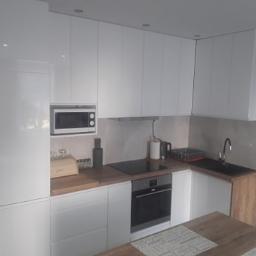 Paweł Naróg Meble - Biała, nowoczesna zabudowa kuchenna z błyszczącymi frontami, drewnianym blatem i wbudowanym sprzętem AGD, takim jak piekarnik i mikrofalówka. Widoczny fragment drewnianego stołu.