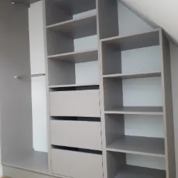 Szara szafa wnękowa z półkami i szufladami, dopasowana do skosu dachu, widoczna podłoga z paneli.