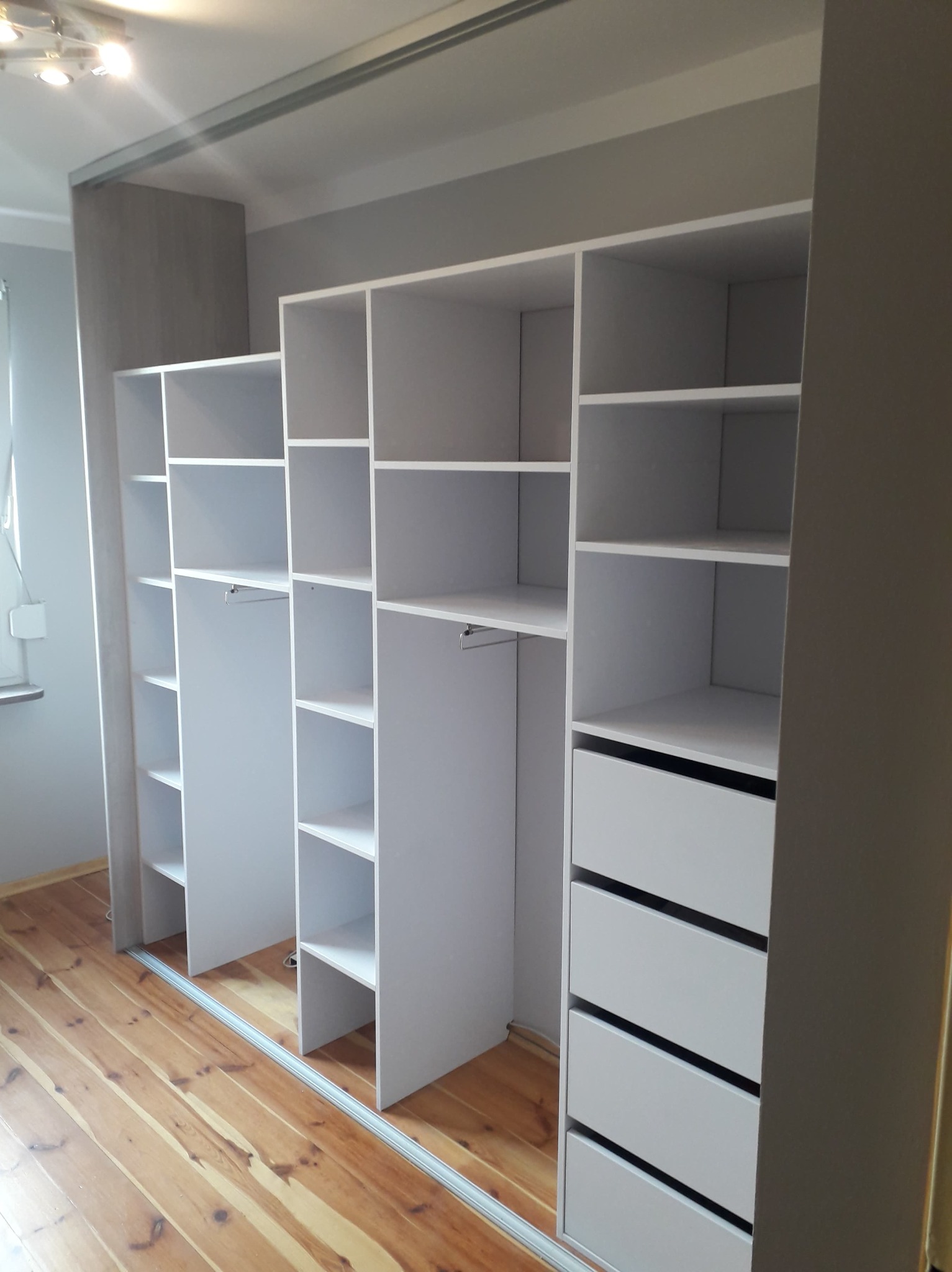 Biała szafa wnękowa z przesuwnymi drzwiami i szufladami, widok na drewnianą podłogę, oświetlenie sufitowe w tle.