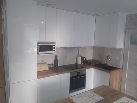 Biała, nowoczesna zabudowa kuchenna z błyszczącymi frontami, drewnianym blatem i wbudowanym sprzętem AGD, takim jak piekarnik i mikrofalówka. Widoczny fragment drewnianego stołu.