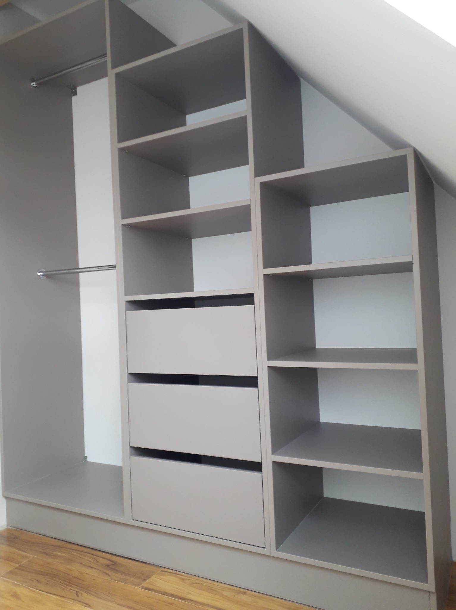 Szara szafa wnękowa z półkami i szufladami, dopasowana do skosu dachu, widoczna podłoga z paneli.