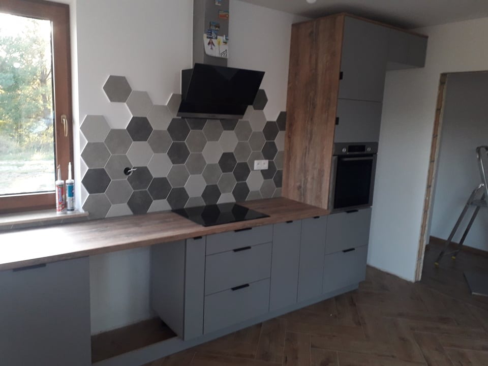 Szare szafki kuchenne z drewnianym blatem i heksagonalnymi płytkami na ścianie, czarny okap i płyta indukcyjna, widoczne silikony przy oknie.