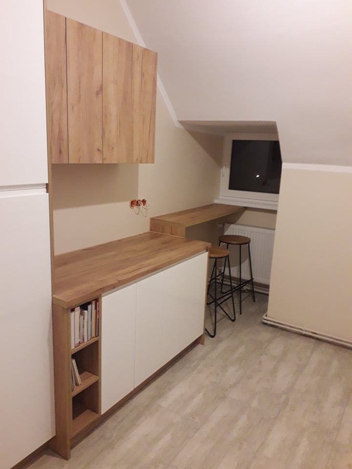 Nowoczesna kuchnia w skosach poddasza: białe fronty szafek dolnych z blatem drewnopodobnym, wiszące szafki górne z forniru drewnopodobnego, drewniany blat roboczy pod oknem, dwa metalowe hokery.