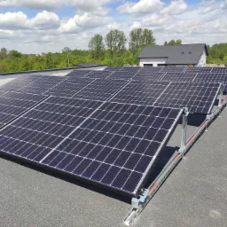 Rząd paneli słonecznych zamontowanych na płaskim dachu z widocznymi elementami montażowymi, w tle budynek mieszkalny i zieleń.