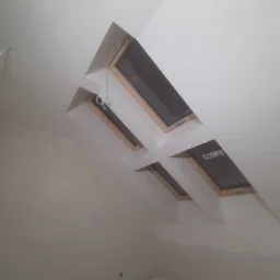 Poddasze po remoncie z trzema oknami dachowymi w drewnianych ramach, świeżo pomalowane na biało, widoczne wystające kable elektryczne.