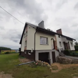 Dwukondygnacyjny dom jednorodzinny z jasną elewacją i ciemnobrązowym cokołem, widoczny z perspektywy niskiego kąta, z zielonym trawnikiem i pochmurnym niebem w tle.